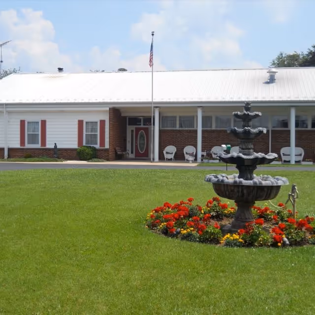 Front exterior of a single-story senior living building with a lawn, flowerbed and tiered fountain in the foreground and porch chairs by the entrance.