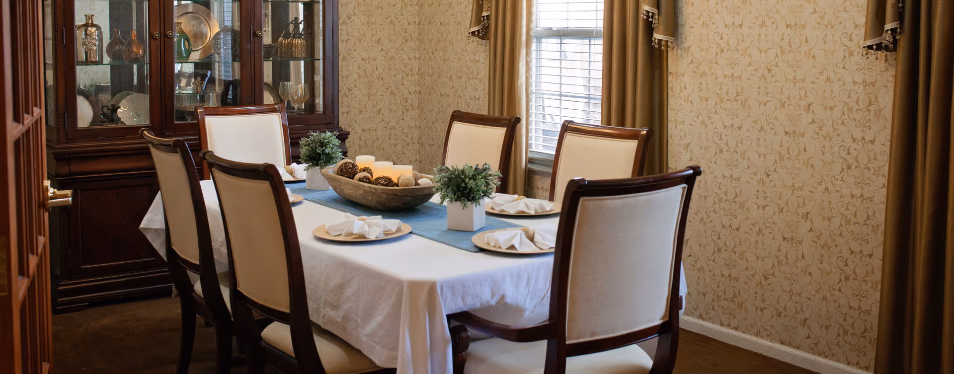 A formal dining room with a rectangular table covered with a white tablecloth and a blue runner. The table is set with six chairs upholstered in cream fabric with dark wood frames. On the table are decorative plants, a wooden bowl with candles and decorative balls, and neatly folded white napkins on plates. In the background, there is a glass-fronted wooden china cabinet displaying various decorative items. The walls have a patterned wallpaper and there is a window with gold curtains.