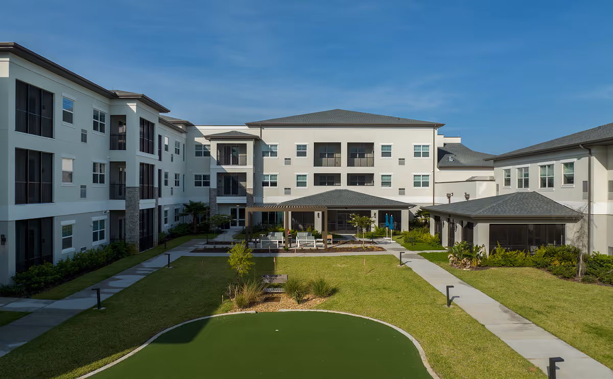 Outdoor courtyard area of a senior living facility with a putting green in the foreground, surrounded by well-maintained grass and pathways. The courtyard is enclosed by a three-story building with balconies and large windows under a clear blue sky.