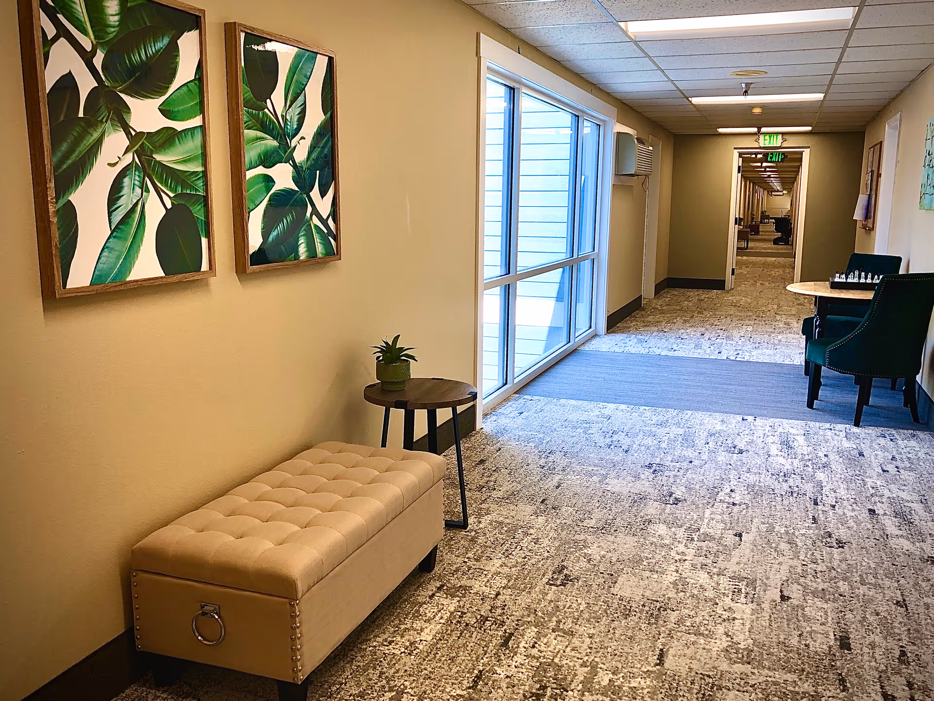 A well-lit hallway in a senior living facility with beige walls and patterned carpet. On the left side, there is a beige tufted bench with a small round wooden table holding a small potted plant. Above the bench, two framed pictures of green leaves hang on the wall. On the right side, there is a small round table with two green upholstered chairs and a chessboard on the table. Large windows on the left side let in natural light, and the hallway extends into the distance with multiple doors and exit signs visible.