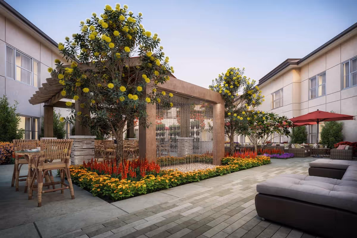 Outdoor courtyard area at Varenita Of Simi Valley featuring a pergola with a water curtain, surrounded by vibrant flower beds and trees with yellow blossoms. There are wicker chairs and tables on the left side and cushioned outdoor seating with a red umbrella on the right side, all set against the backdrop of the facility building.