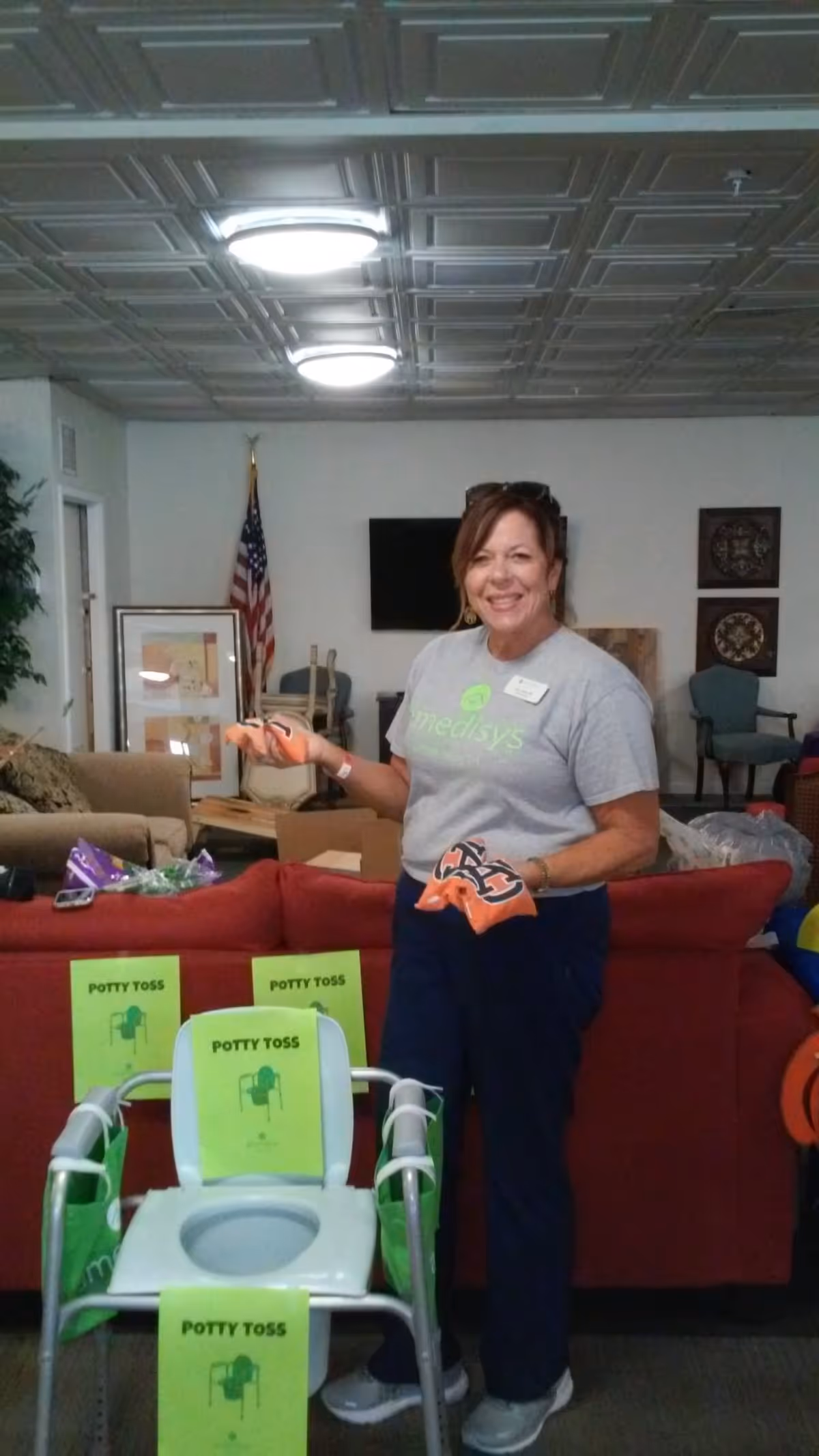 A smiling woman wearing a gray Medisys t-shirt and dark pants stands in a living room area holding orange bean bags. In front of her is a chair with a toilet seat and several green signs labeled 'POTTY TOSS'. The room has a red couch, an American flag, framed pictures, and a TV mounted on the wall.