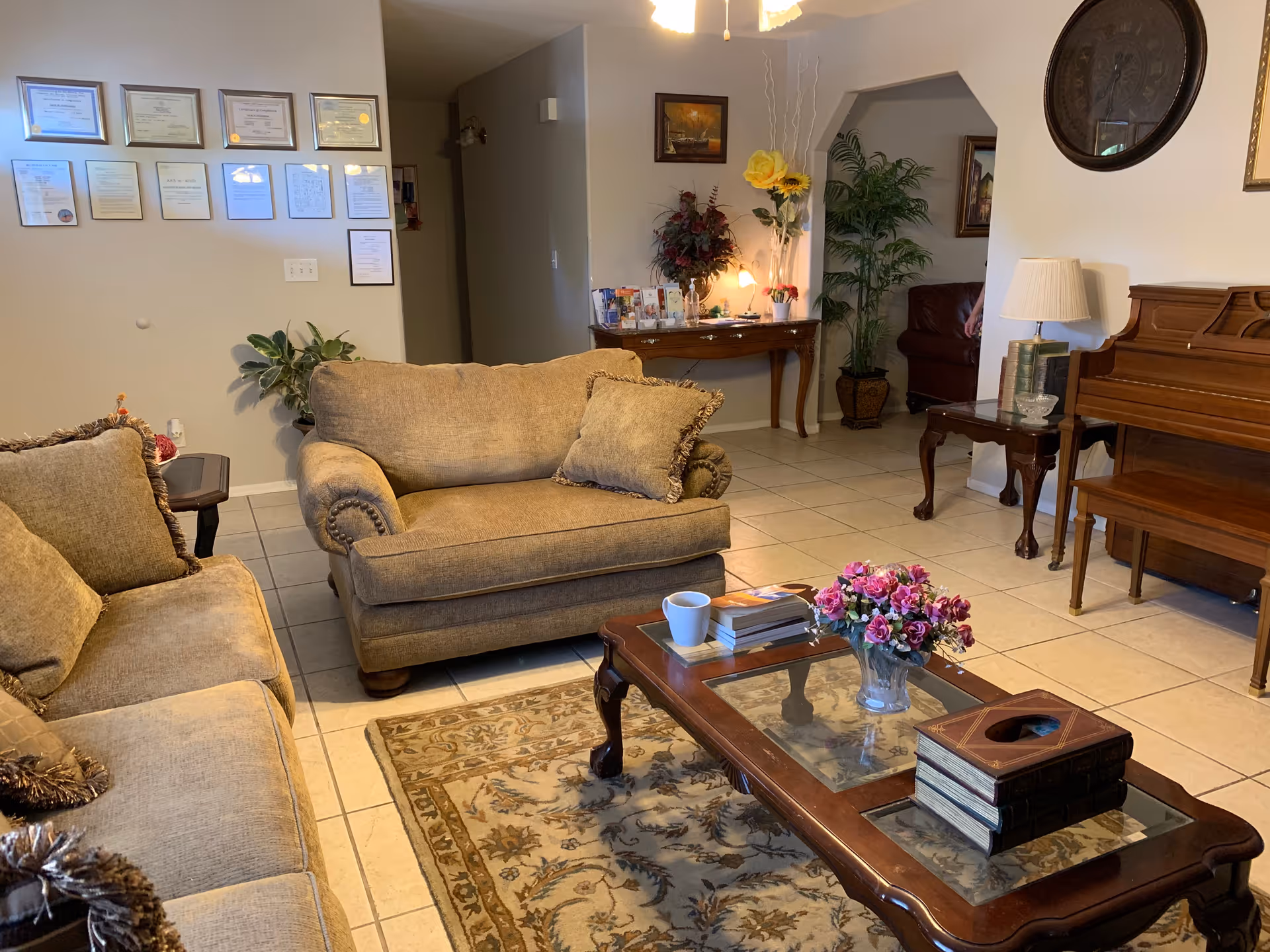 Comfortable living room with upholstered sofas, a glass-top coffee table with flowers and books, a piano, and framed certificates on the wall.