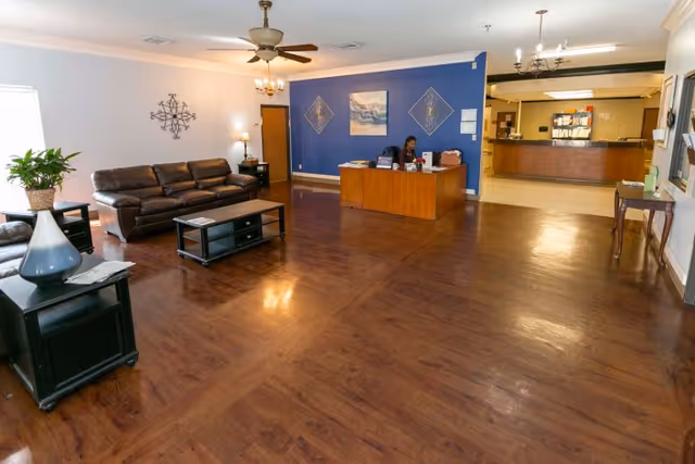 A spacious reception area with wooden flooring, featuring a brown leather sofa and matching armchair, a coffee table, and a side table with a plant. A receptionist is seated at a wooden desk against a blue accent wall decorated with artwork. In the background, there is a reception counter with shelves and files. The room is lit by ceiling fans with lights and wall sconces.