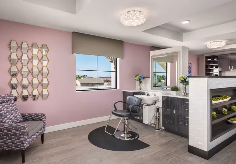 A modern salon area inside a senior living facility with a black salon chair in front of a white sink and a large mirror. The walls are painted light purple, and there is a window with a view of rooftops outside. A patterned armchair is placed against the wall with a decorative honeycomb mirror above it. The space is well-lit with ceiling lights and decorated with small plants and flowers.