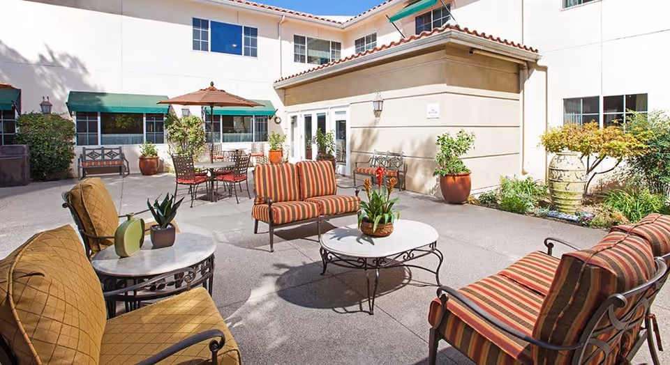 Outdoor patio area at Atria Santa Clarita featuring cushioned metal chairs and sofas with striped and solid yellow cushions, round tables with potted plants, a dining table with an umbrella, and surrounding greenery and potted plants against a beige building with green awnings.