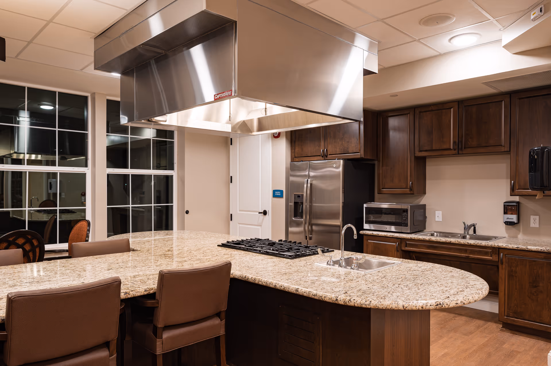 Modern kitchen area with a large granite island countertop featuring a built-in gas stove and sink. The kitchen has dark wooden cabinets, a stainless steel refrigerator, a microwave, and a stainless steel range hood above the stove. There are several brown cushioned chairs around the island, and large windows in the background.