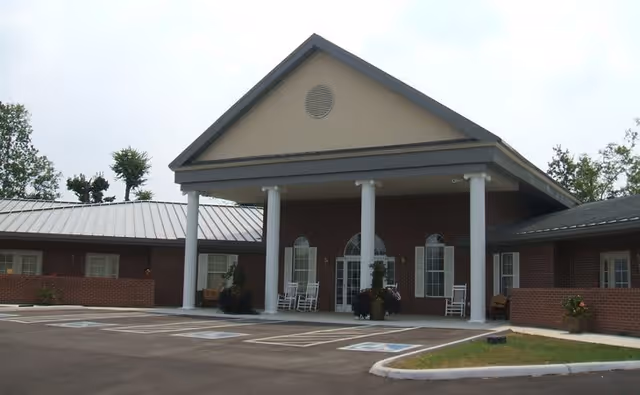 Front exterior view of Trousdale Senior Living Center showing a brick building with a covered entrance supported by white columns, several rocking chairs on the porch, and a parking area with marked spaces including handicapped spots.