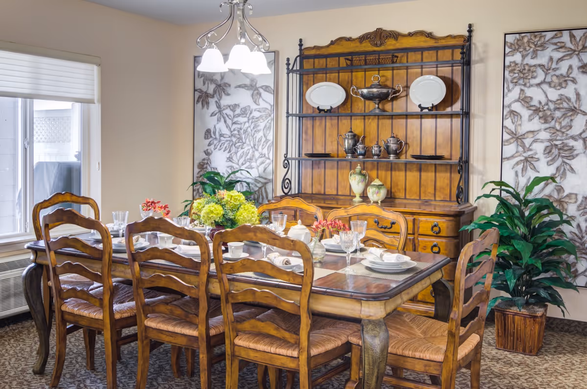 A well-lit dining room with a wooden table set for a meal, matching chairs, a decorative hutch and potted plants.