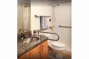 A small accessible bathroom with a granite-top sink, toilet, grab bars, and a shower with curtain.