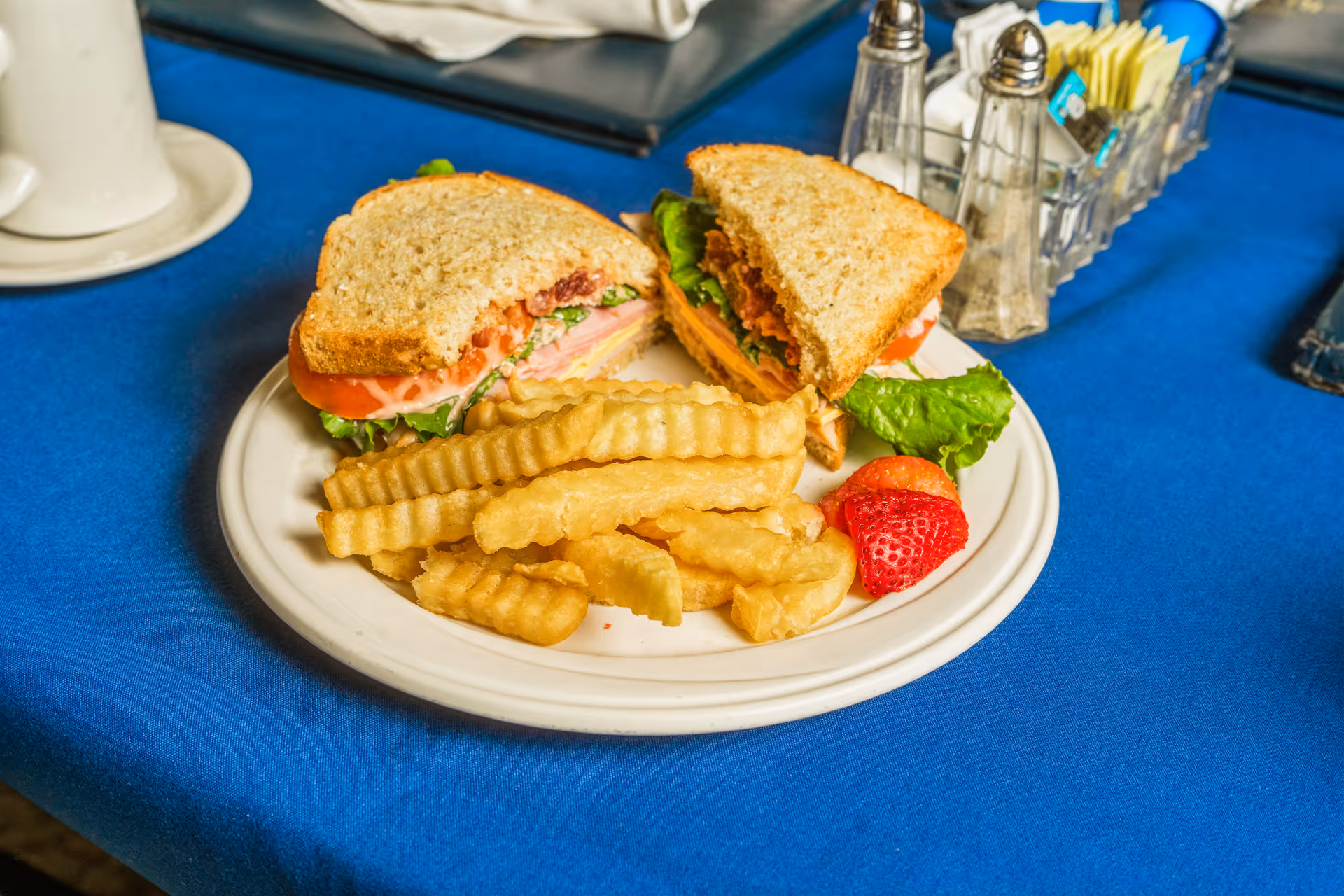 A plate with a halved sandwich, crinkle-cut fries, and a strawberry on a blue tablecloth with salt, pepper and sugar packets nearby.
