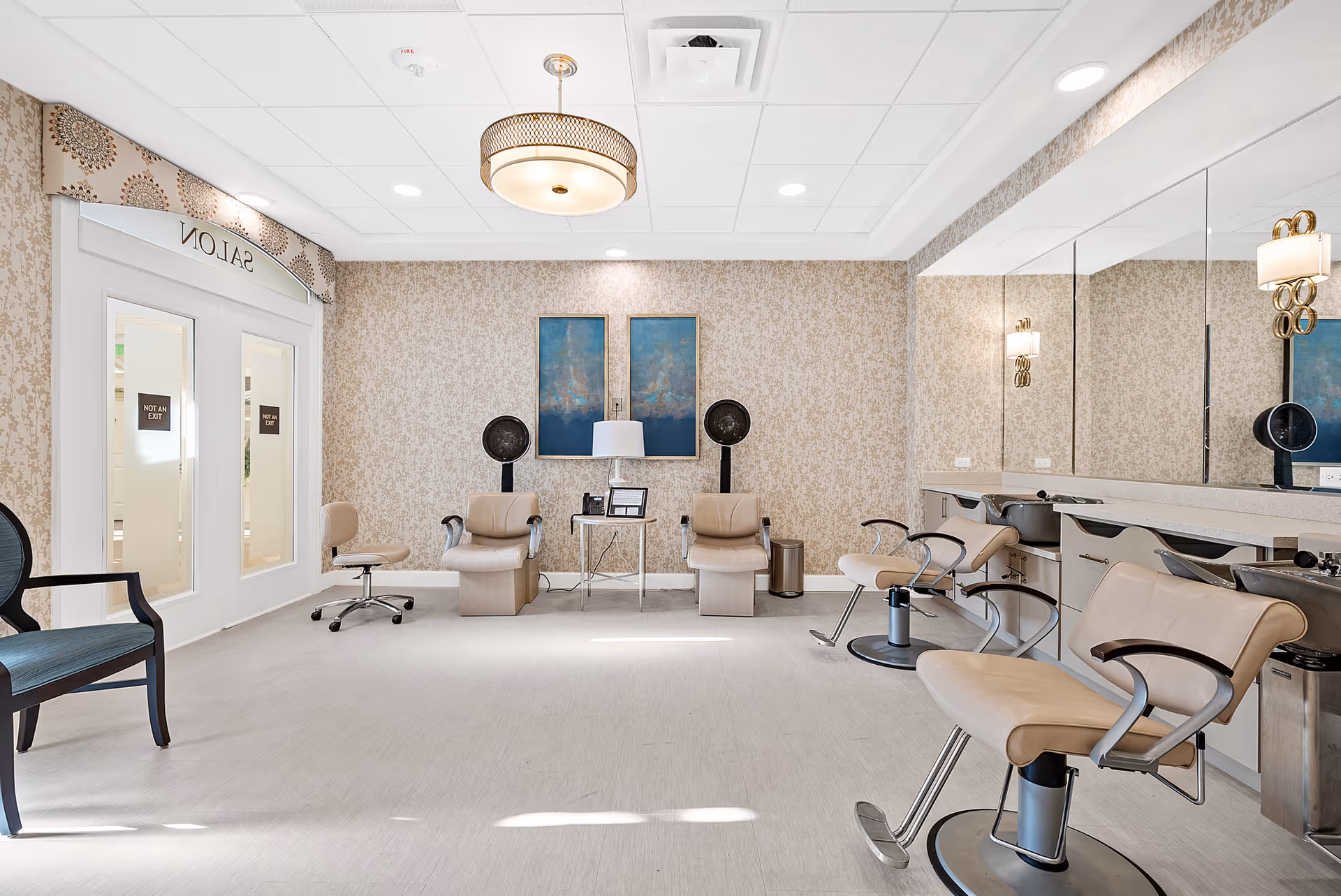 Interior view of a salon with beige walls and flooring, featuring salon chairs, hair dryers, a large mirror on one wall, and a decorative ceiling light fixture. There are two framed artworks on the back wall and a door labeled 'SALON' with 'NOT AN EXIT' signs on the glass panels.