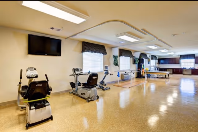A bright rehabilitation exercise room with recumbent bikes, therapy equipment and parallel bars, with a kitchenette area in the background.