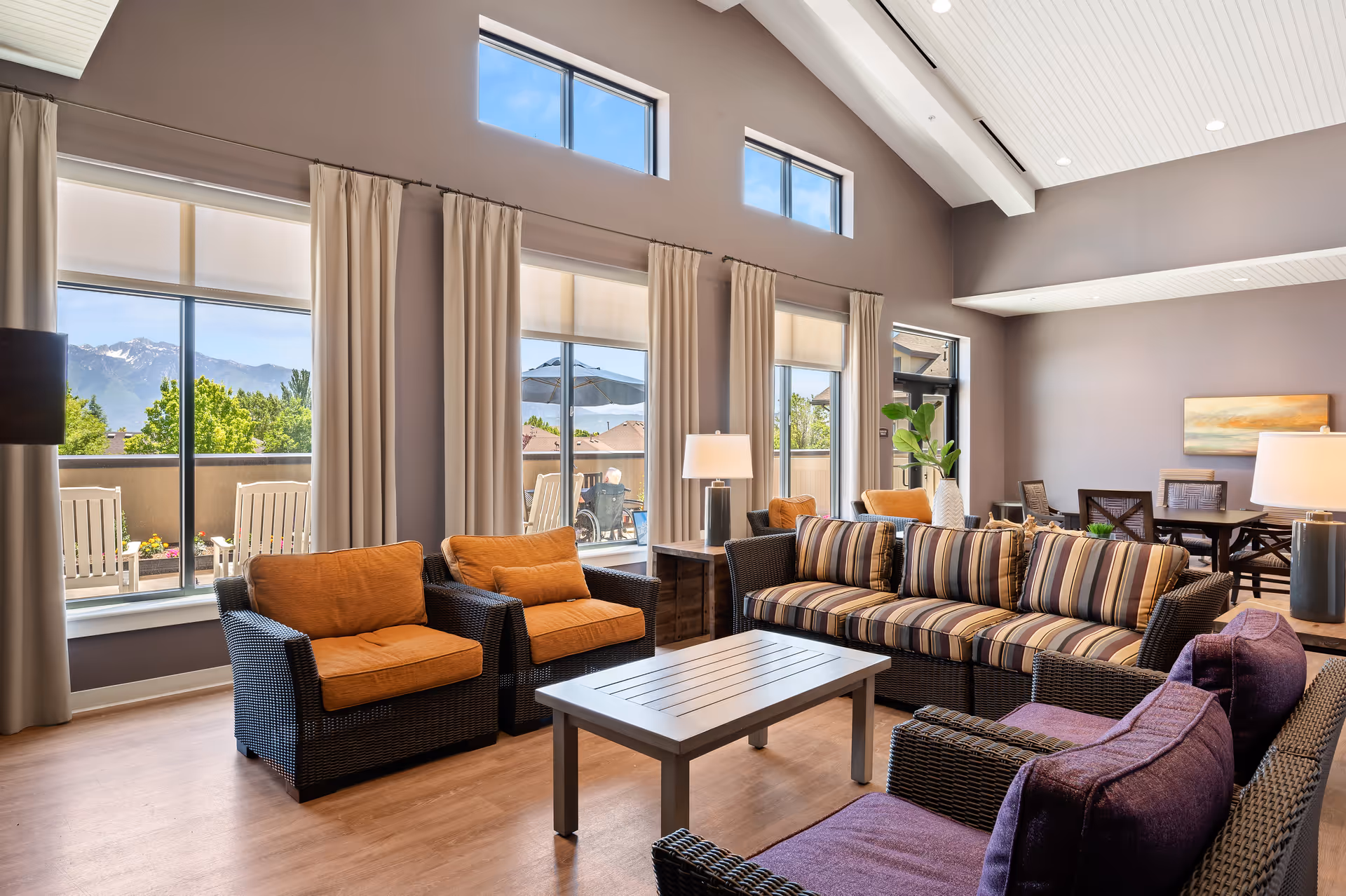 Bright communal living room with wicker sofas and chairs around a coffee table, large windows, and high vaulted ceilings.