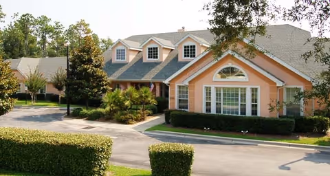 Exterior view of a peach-colored building with a gray roof, multiple windows including dormer windows, surrounded by greenery and trees, with a paved driveway and trimmed hedges in front.
