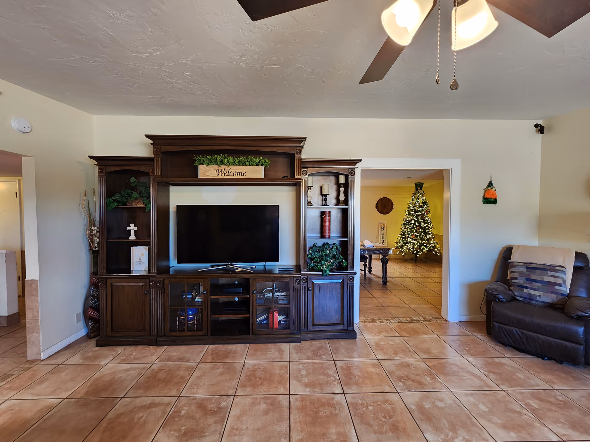 A living room with a large dark wooden entertainment center holding a flat-screen TV, decorative plants, candles, and a 'Welcome' sign. To the right, there is a brown leather recliner with a patterned pillow. Through an open doorway, a decorated Christmas tree and a pool table are visible in an adjacent room. The floor is tiled, and a ceiling fan with lights is partially visible at the top right.