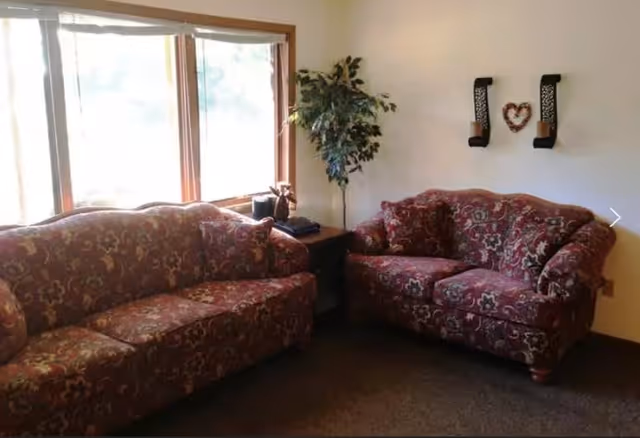 A cozy living room area with two patterned red sofas facing each other, a small wooden side table between them holding a plant and some items, a large window letting in natural light, a potted plant in the corner, and two wall-mounted candle holders with a heart decoration between them.
