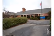 Exterior view of a single-story brick building with a gray roof, a chimney, and an American flag on a flagpole in front. There are bushes and a paved driveway leading to the entrance, with a blue sign near the door.