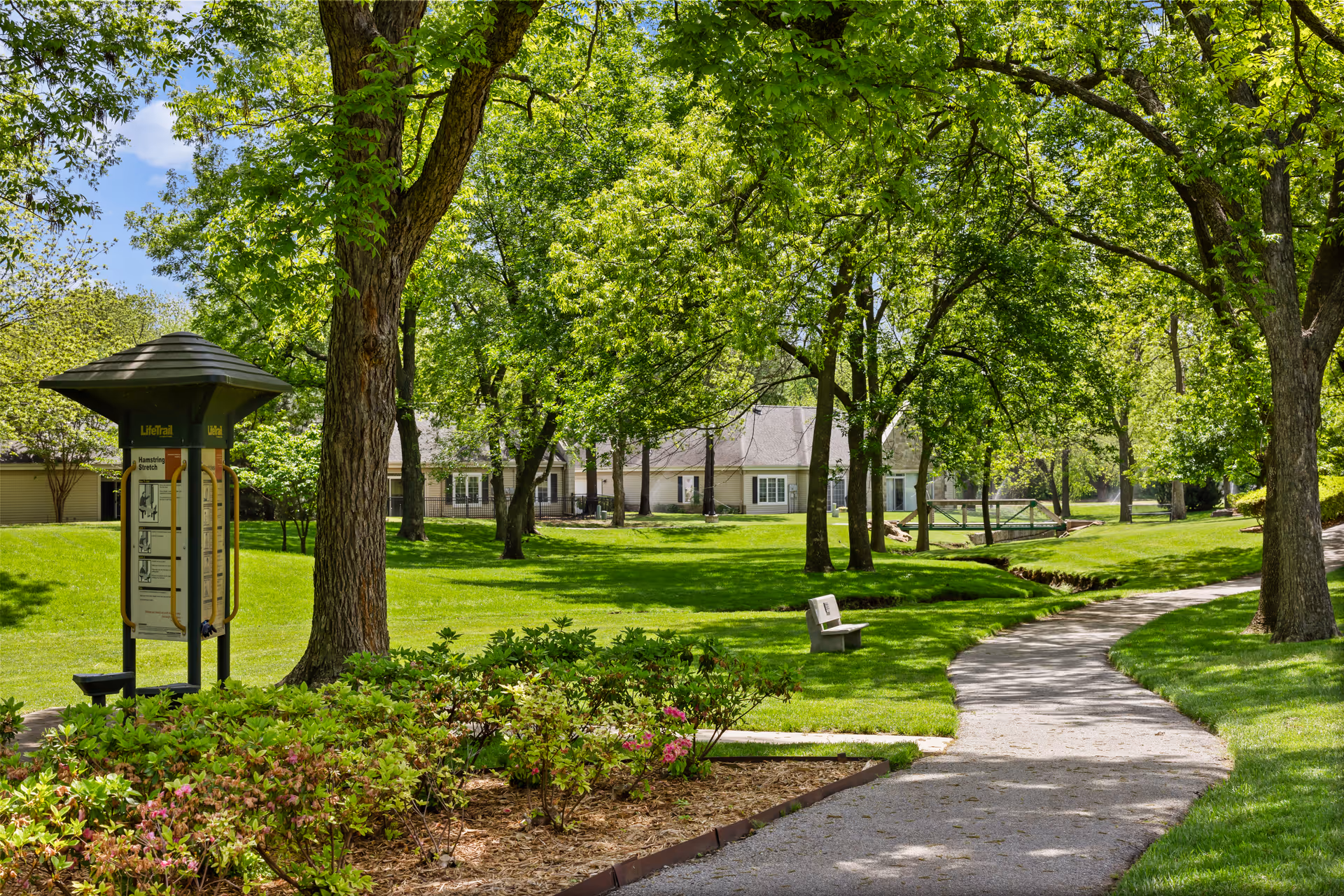 Tree-lined grassy courtyard with a winding path, bench, exercise kiosk, and buildings in the background.