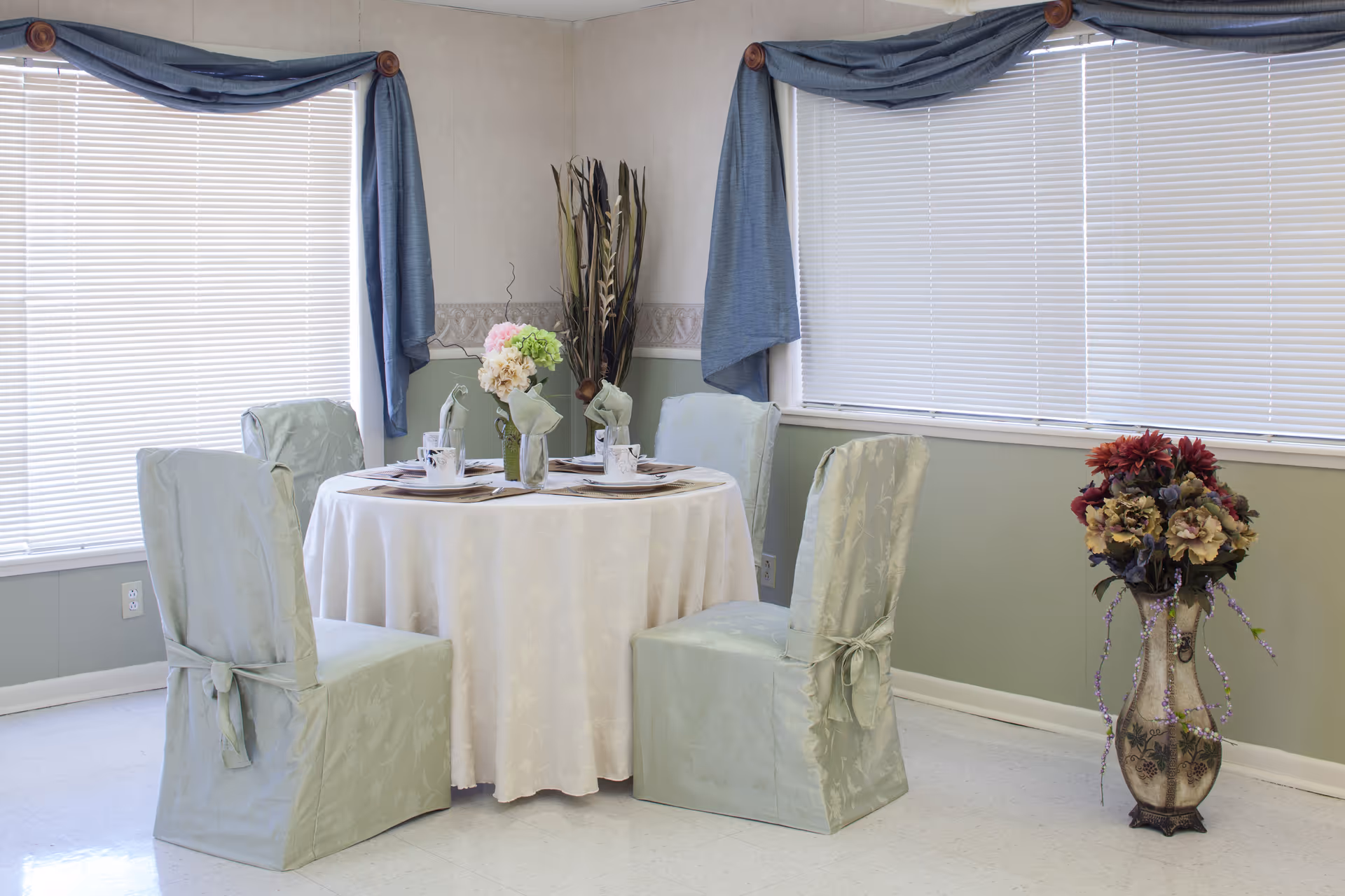 Round dining table set with four skirted chairs and floral centerpieces in a bright room with large blinds and blue valances.