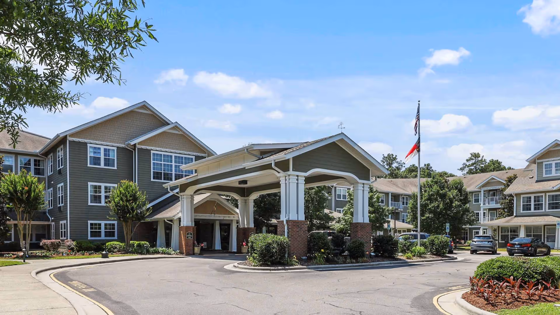 Front entrance of a senior living facility with a covered porte-cochere, driveway, flagpoles, and landscaped grounds.