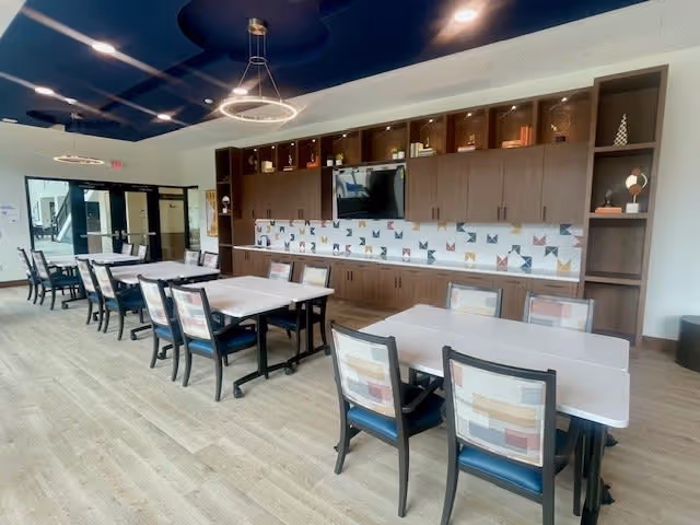 A spacious dining area with multiple tables and chairs arranged in rows. The room features a long wooden cabinet with upper and lower storage, a mounted flat-screen TV, and a decorative backsplash with a colorful geometric pattern. Modern circular pendant lights hang from the ceiling, and large windows and glass doors provide natural light.
