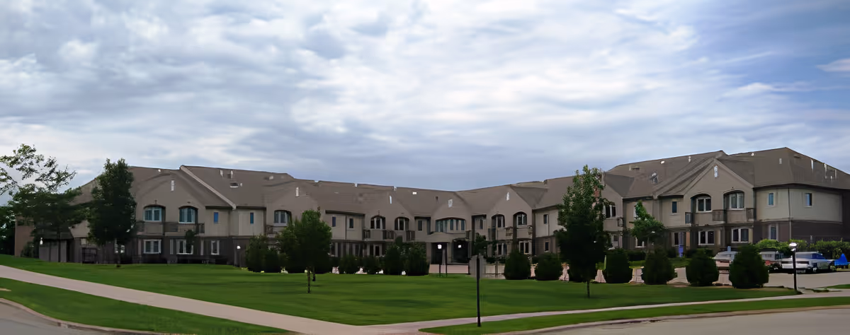 Wide exterior view of a large, multi-story senior living facility building with a sloped roof, surrounded by green lawns, trees, and a paved walkway under a cloudy sky.