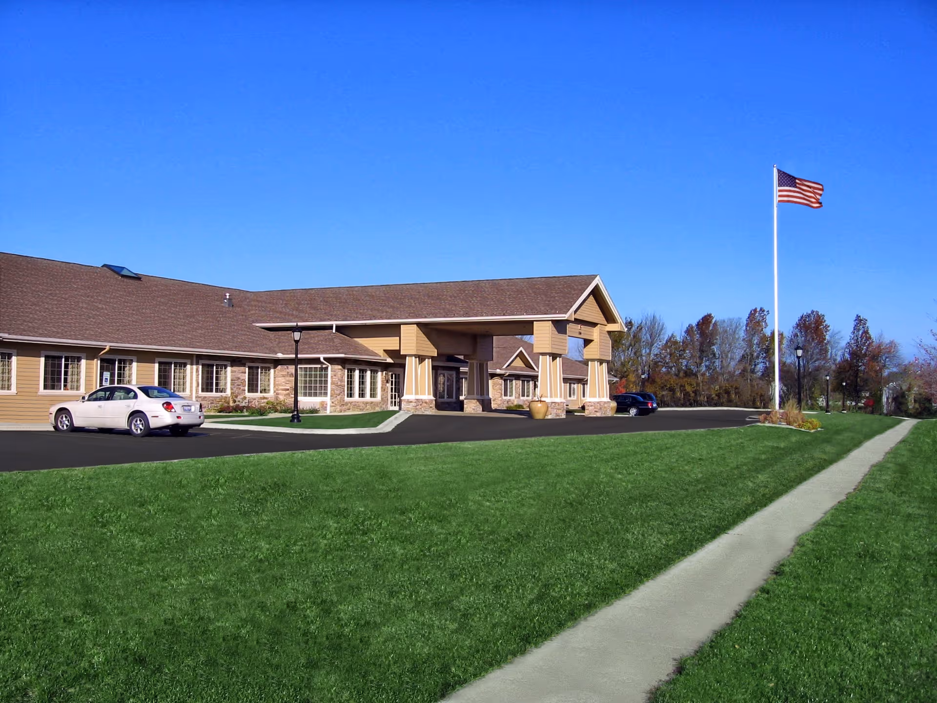 Exterior view of Amber Glen Memory Care facility showing a single-story building with a covered entrance, a parking lot with a few cars, a green lawn, a sidewalk, and an American flag on a tall flagpole against a clear blue sky.