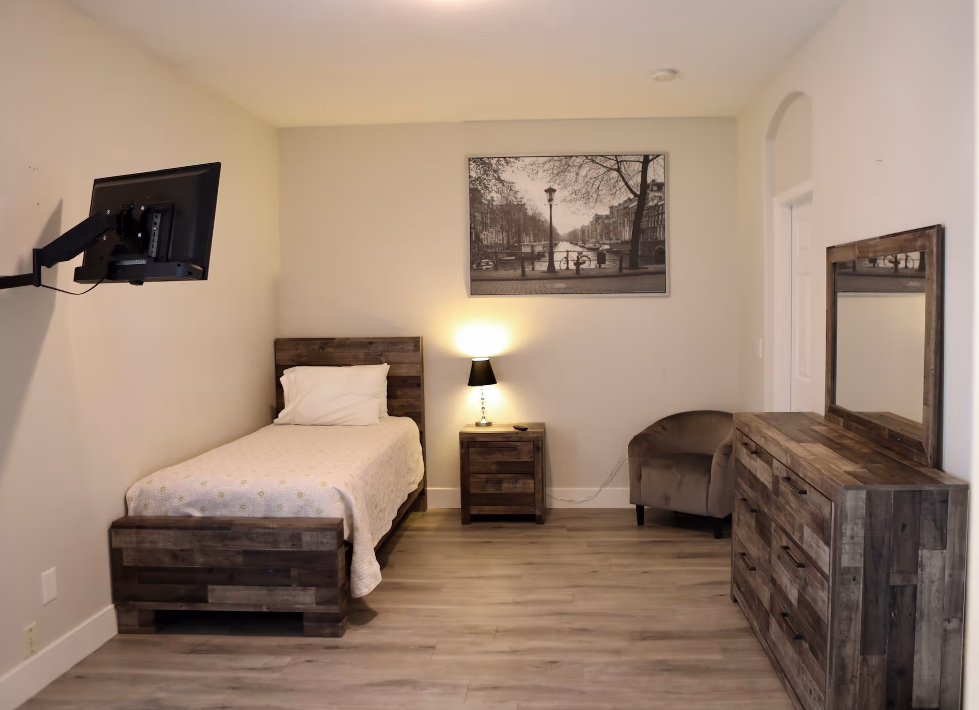 A simple bedroom with a single wooden bed with white bedding, a wooden nightstand with a lamp, a wooden dresser with a mirror, a brown armchair, and a wall-mounted TV. A black and white framed picture hangs above the nightstand.