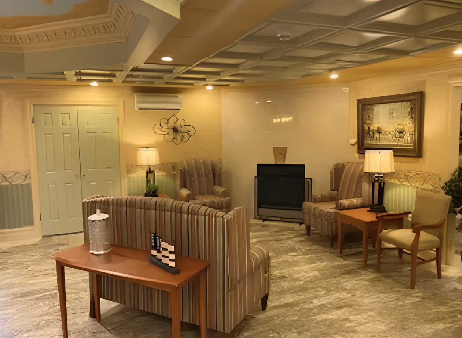 A cozy living room area with striped upholstered chairs and a sofa arranged around a fireplace. There are two wooden side tables with lamps and decorative items, a framed painting on the wall, and soft lighting creating a warm ambiance.