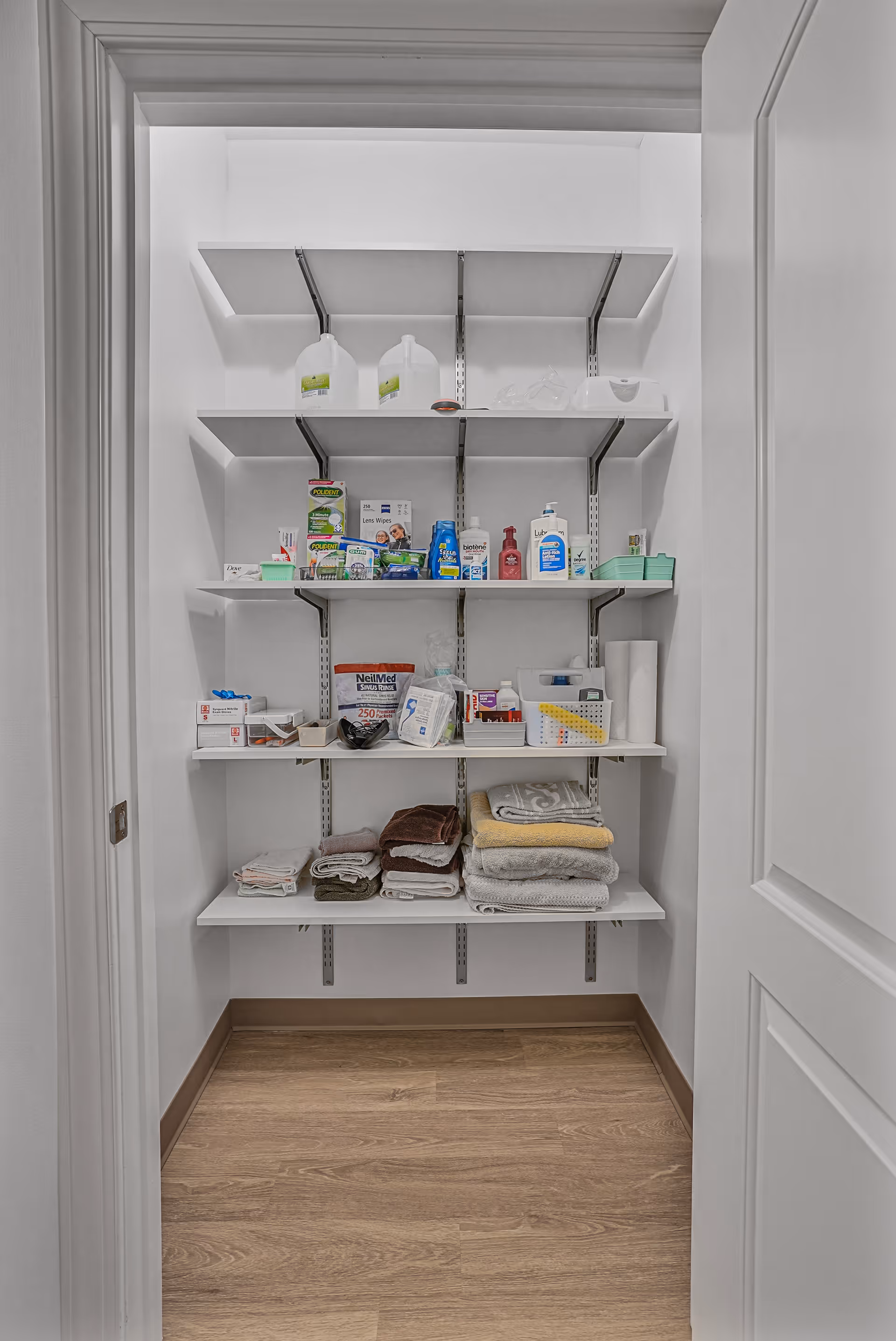 A small storage closet with white shelves holding various household items including folded towels, cleaning supplies, paper towels, and personal care products. The closet has white walls and a wooden floor.