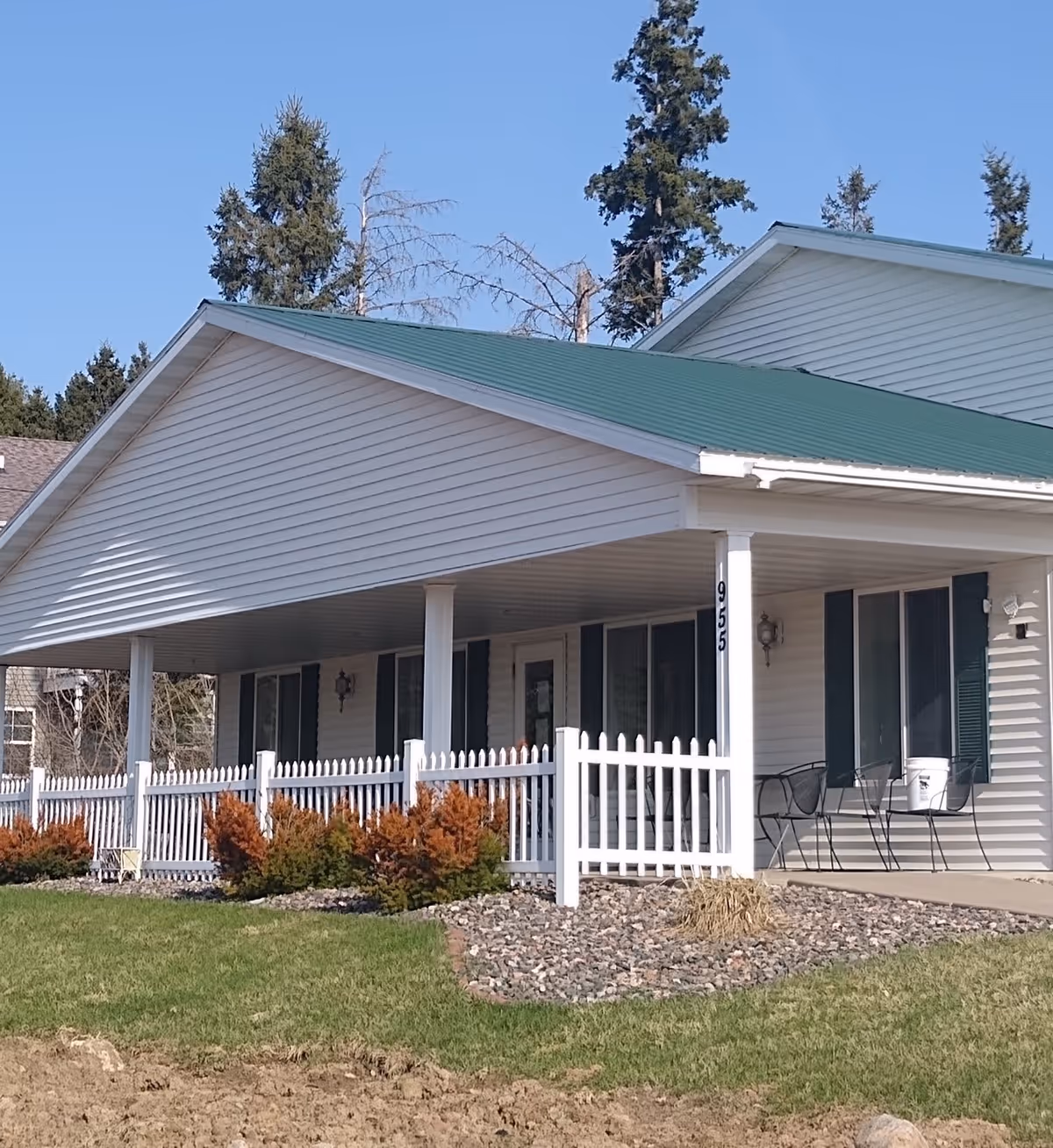 Exterior view of a single-story building with white siding and a green metal roof. The building has a covered porch supported by white columns, a white picket fence, and some shrubs in front. There are two black metal chairs and a white bucket on the porch. The building number 955 is visible on one of the columns. Tall evergreen trees are in the background under a clear blue sky.