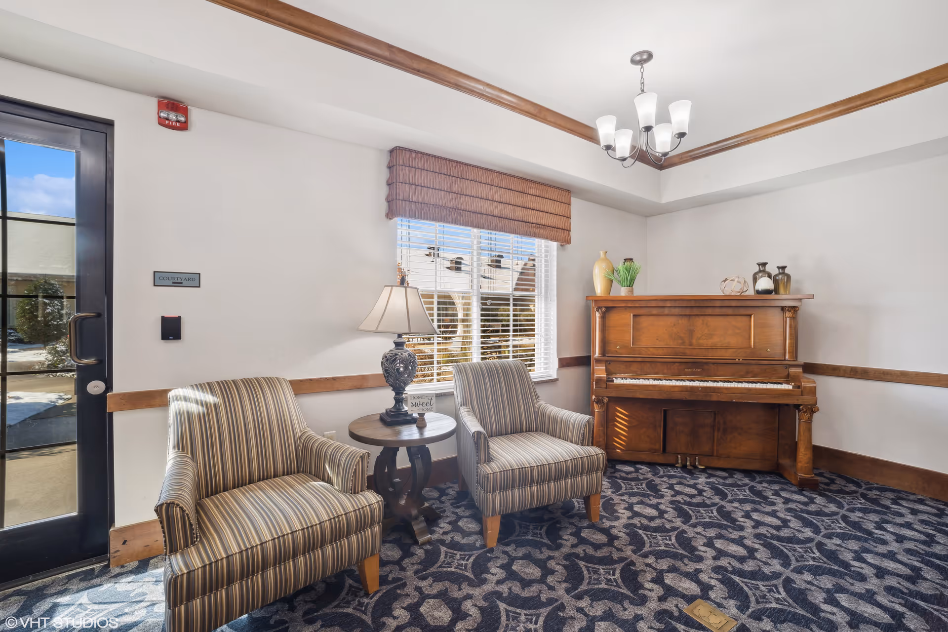 A cozy interior room with two striped armchairs separated by a round wooden side table with a decorative lamp and a small sign that reads 'Home Sweet Home'. Behind the chairs is a window with blinds and a brown valance. To the right, there is a wooden upright piano adorned with decorative vases and ornaments. The room features a patterned carpet, white walls with wooden trim, and a ceiling light fixture. A glass door labeled 'Courtyard' is visible on the left side.