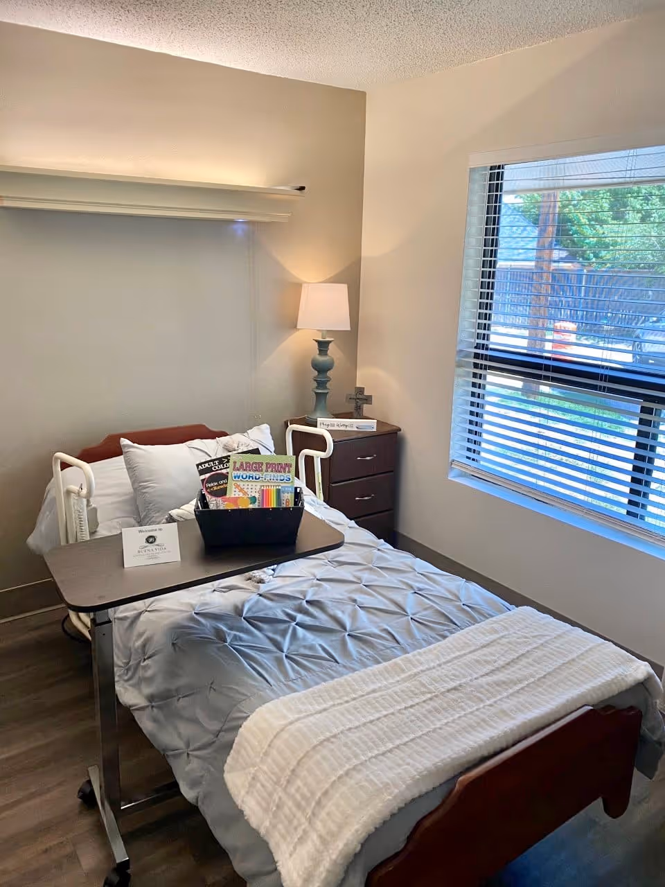 A neatly made single bed in a nursing facility room with a gray comforter and white blanket. A wooden bedside table with a lamp and a small cross is next to the bed. A window with blinds lets in natural light. A tray table on the bed holds a basket with large print word-find books and colored pencils.
