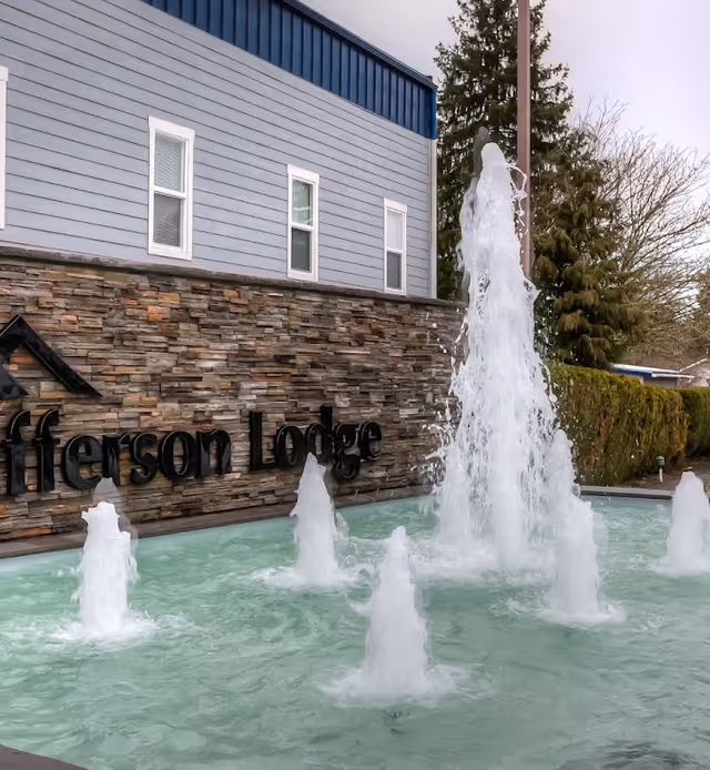 Outdoor water fountain with multiple jets of water shooting upwards in front of a building with a stone facade and blue siding. The building has three windows and a sign that reads 'Jefferson Lodge'. Trees and shrubs are visible in the background.