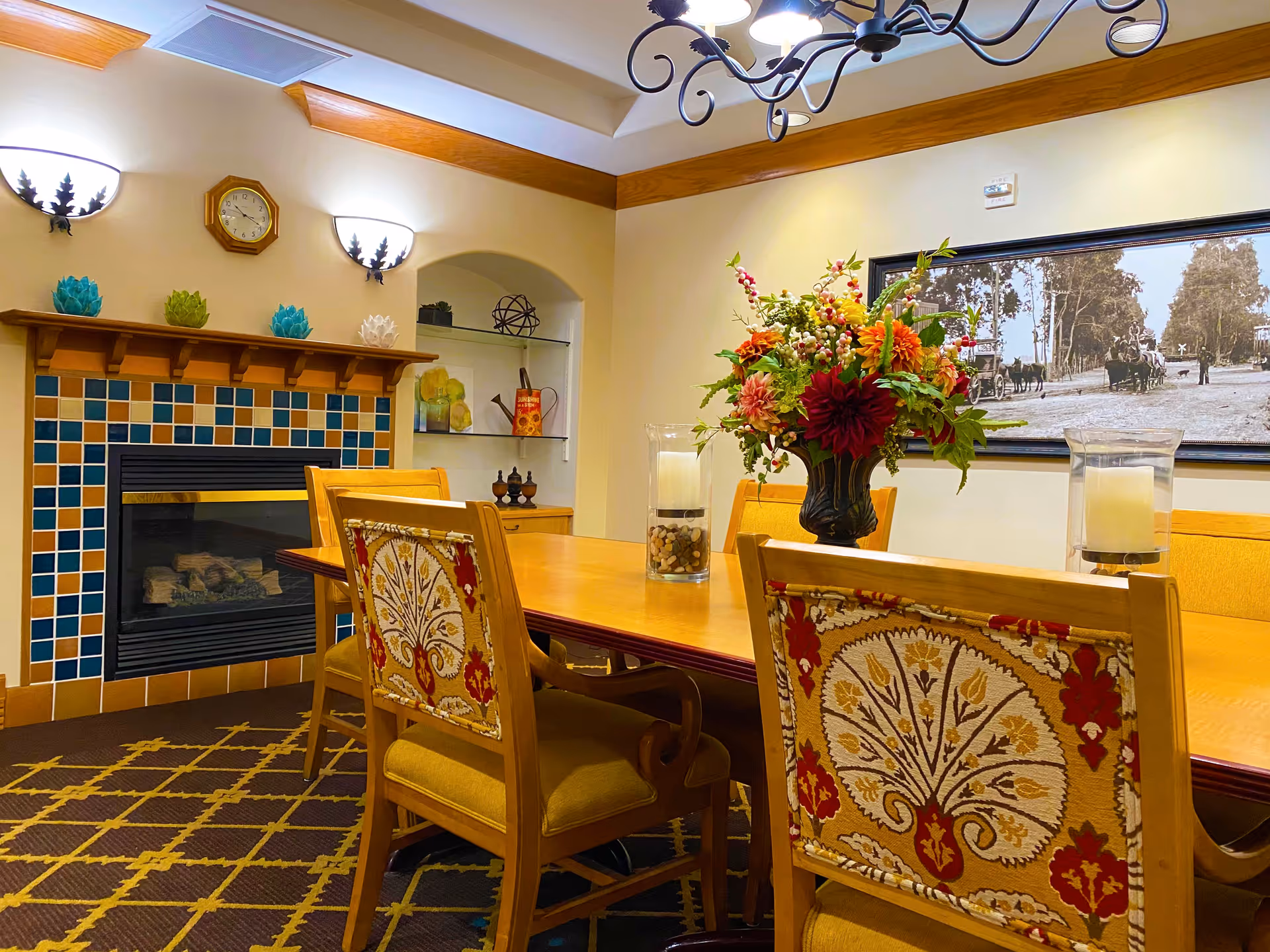 A cozy dining room with a wooden table surrounded by upholstered chairs featuring a floral pattern. The room has a fireplace with a colorful tiled surround and a wooden mantel decorated with small artichoke-shaped ornaments. Above the mantel is a clock and two wall sconces. A large floral arrangement and two glass candle holders are on the table. A black and white framed photograph hangs on the wall, and the room has warm lighting with a chandelier overhead.