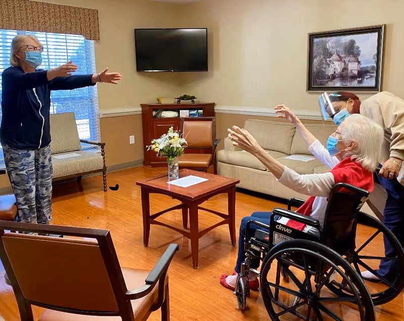Masked residents and a staff member reach toward each other in a seating area with chairs, a coffee table, and a wall-mounted TV.