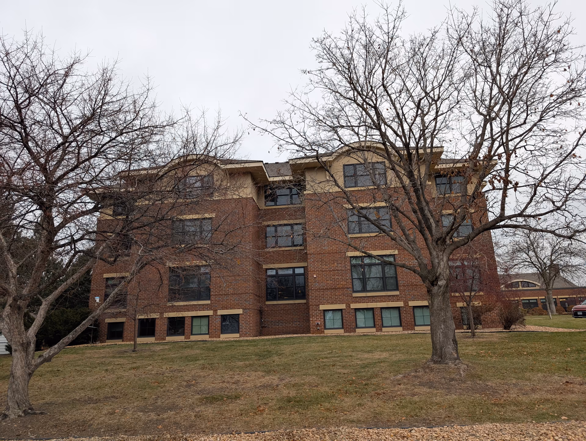 Exterior view of a three-story brick building with multiple windows, surrounded by leafless trees and a grassy area under an overcast sky.