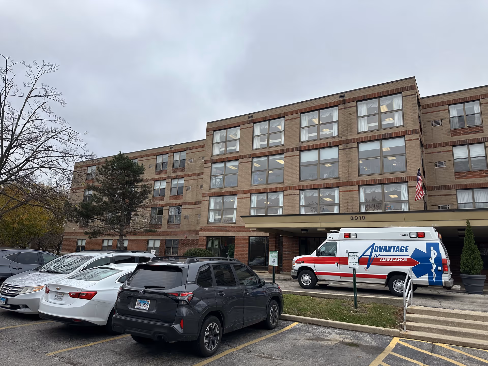 Exterior view of a multi-story brick building with large windows and an American flag near the entrance. Several cars are parked in front, including an ambulance with 'Advantage Ambulance' written on its side. The sky is overcast.