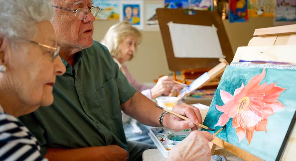 Three elderly individuals engaged in painting at easels, focusing on a close-up of a man and woman painting a large pink flower on a canvas, with another woman painting in the background.