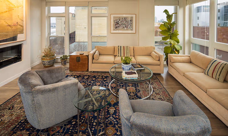 Bright furnished living room with sofas, armchairs, glass coffee tables, a patterned rug and large windows.
