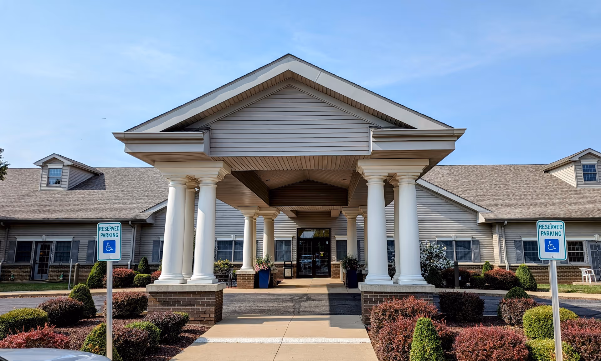 Front entrance of a single-story care facility with a covered portico supported by white columns, accessible parking signs, and landscaped shrubs.