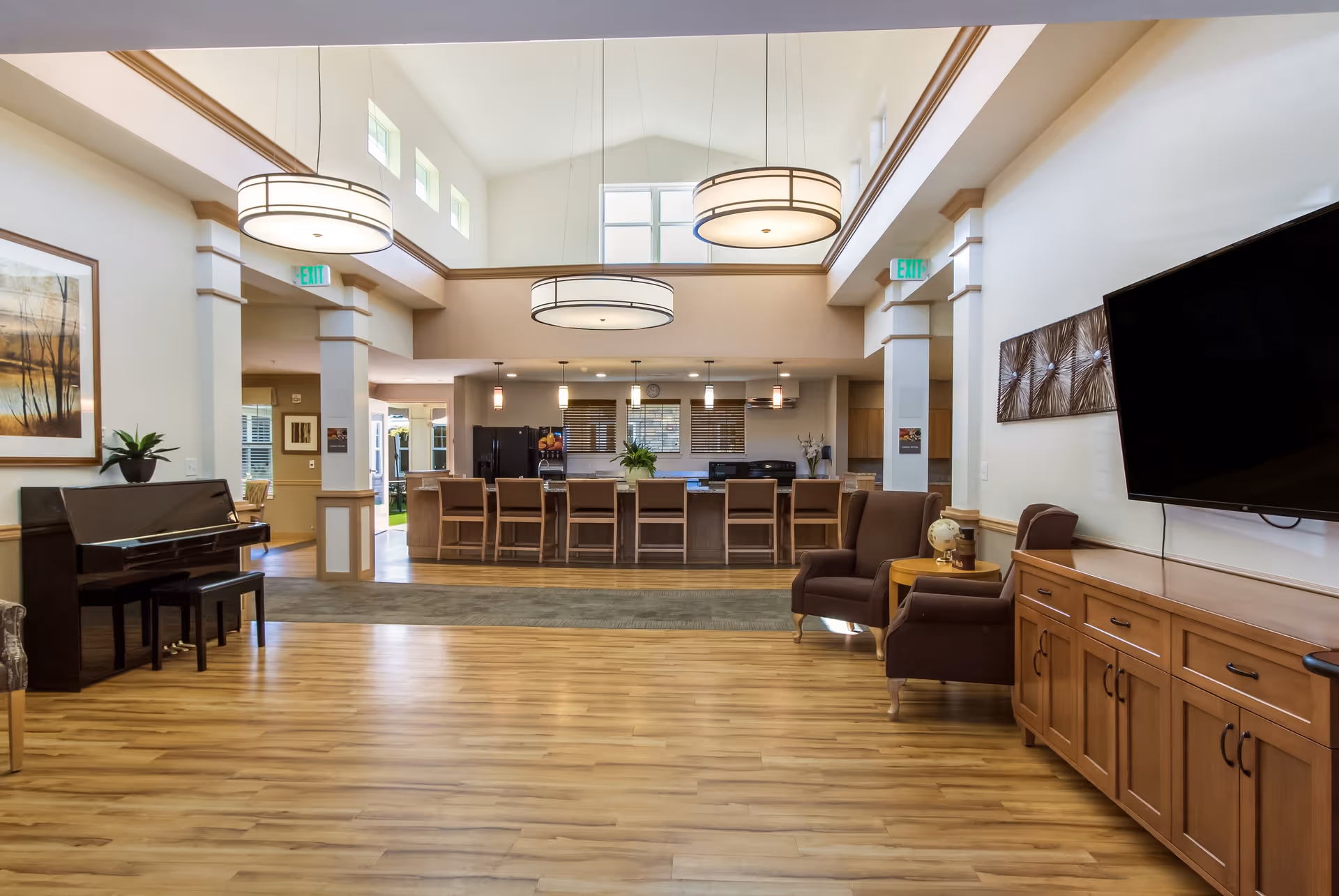 Spacious and well-lit common area in a senior living facility featuring a wooden floor, a piano with a bench on the left, a large kitchen island with multiple chairs in the background, two armchairs with a small round table between them on the right, a large flat-screen TV mounted on the wall above a wooden cabinet, and modern hanging light fixtures from a high ceiling with windows near the top.