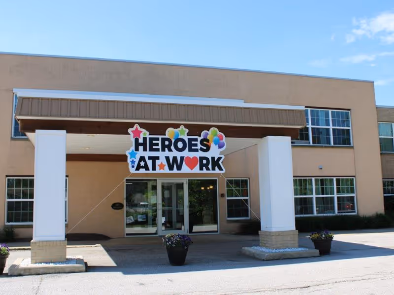 Front exterior view of a beige two-story building with large windows and a covered entrance. A colorful sign hanging from the entrance roof reads 'HEROES AT WORK' with stars, balloons, and a heart symbol. There are flower pots placed near the entrance.