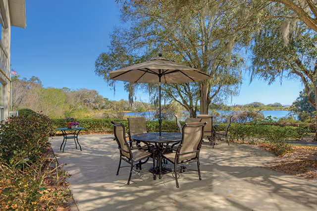 Outdoor patio area with a round table and four chairs under a large umbrella. Additional tables and chairs are visible in the background near a hedge. Trees and a body of water are seen beyond the patio under a clear blue sky.