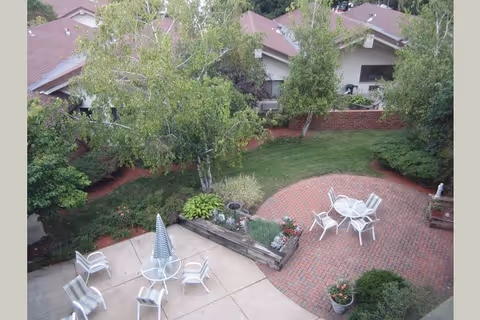 View of an outdoor patio area with white chairs and tables, some with umbrellas, surrounded by greenery including trees, bushes, and a grassy lawn. The patio has a combination of concrete and brick surfaces, and residential-style buildings with brown roofs are visible in the background.