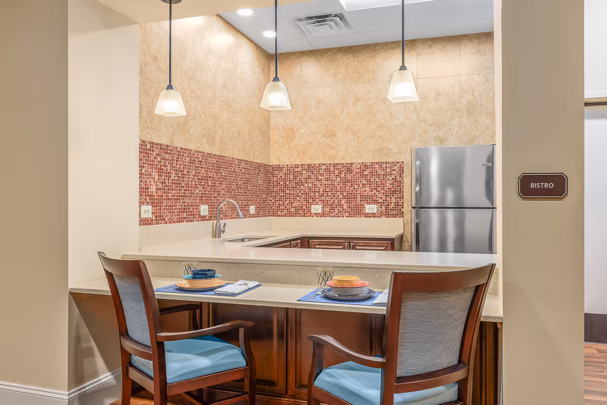 A small kitchen area with a beige countertop and red mosaic tile backsplash. Two wooden chairs with blue cushions are placed at the counter, which is set with plates, bowls, and napkins. Three pendant lights hang from the ceiling, and a stainless steel refrigerator is visible in the background. A sign on the wall reads 'Bistro'.