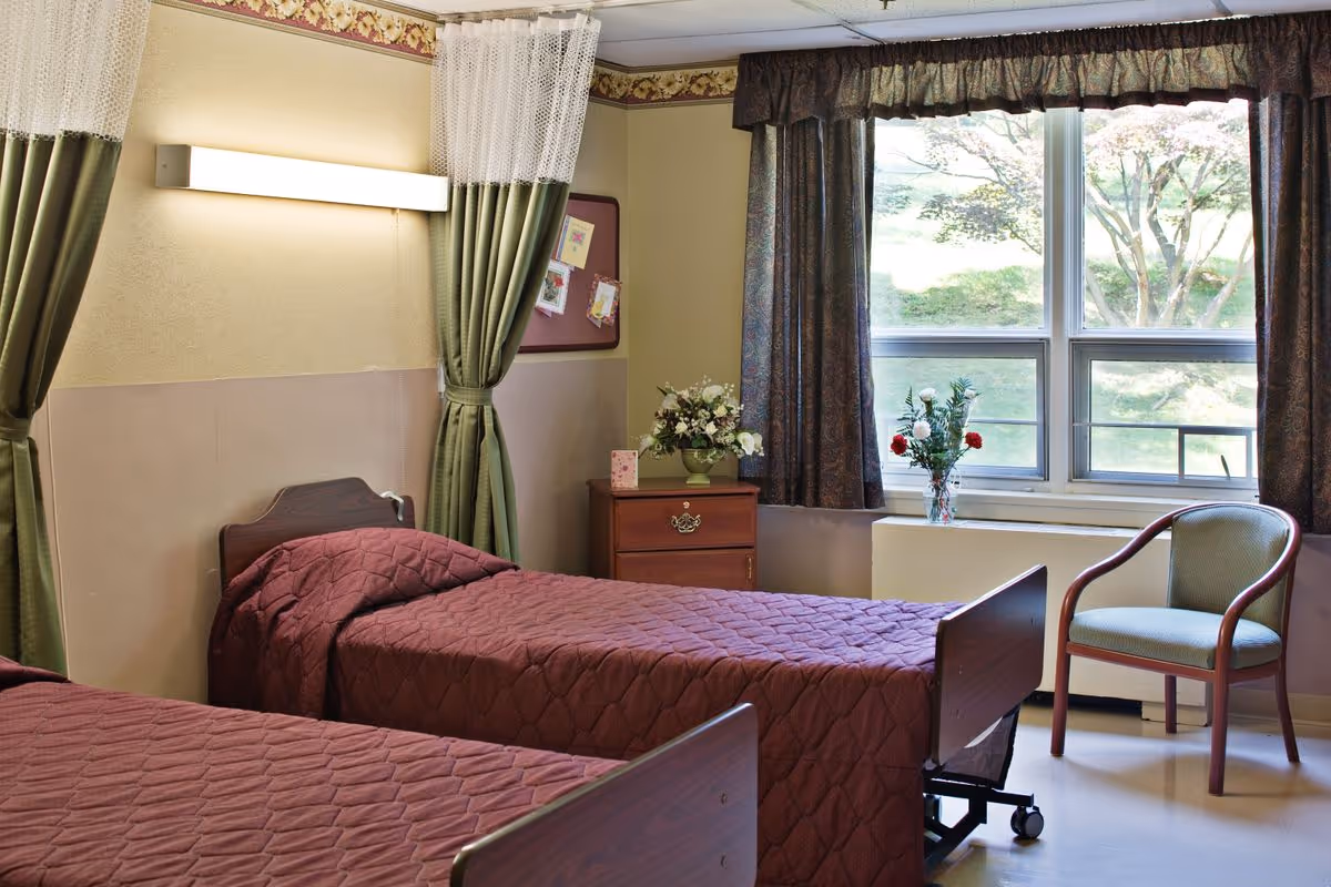 Two twin beds with burgundy quilts in a senior living room with a nightstand, window, and a chair.