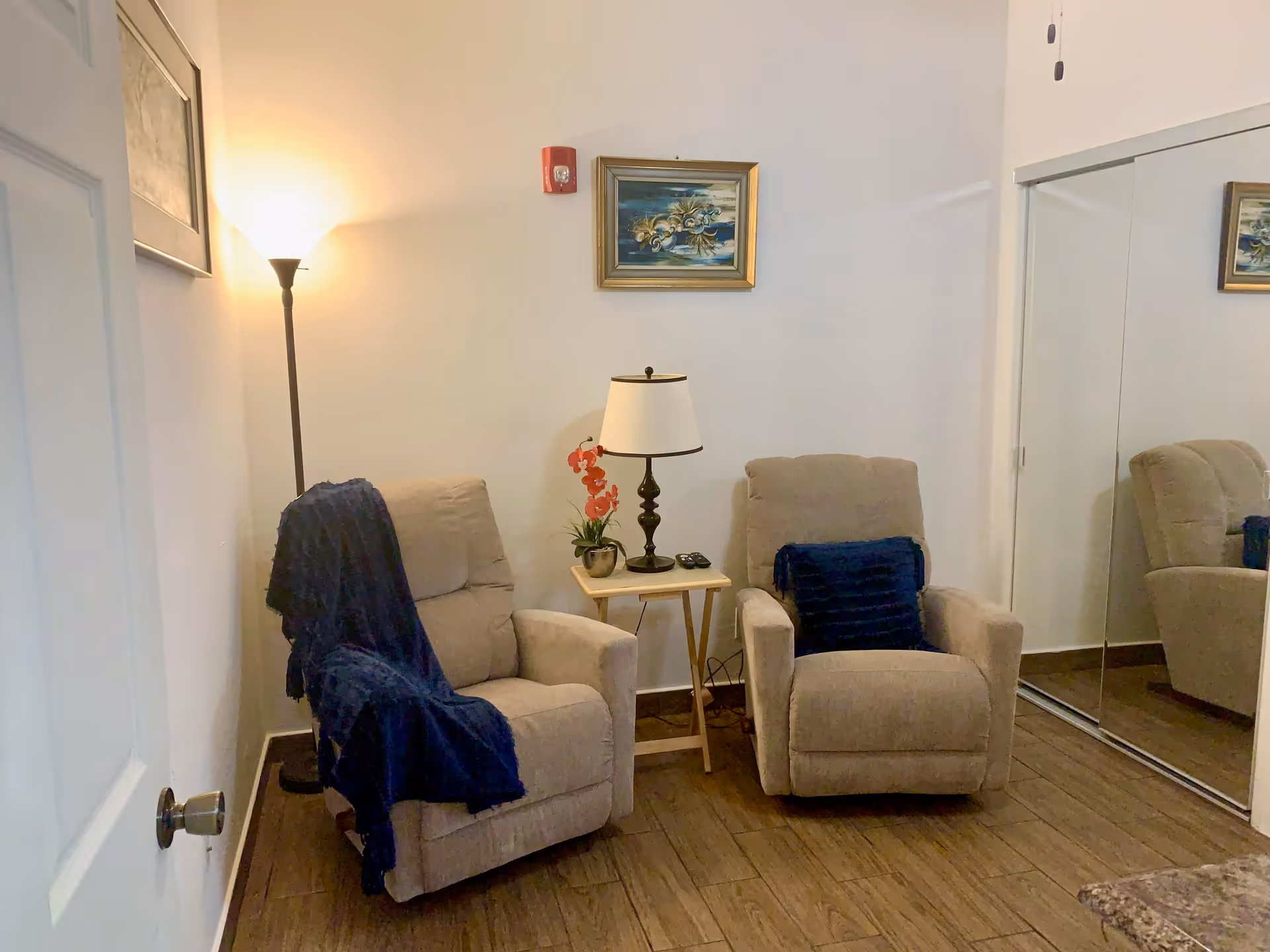 A cozy sitting area with two beige recliner chairs, each adorned with a dark blue throw blanket or pillow. Between the chairs is a small wooden side table with a lamp and a small potted plant with red flowers. A floor lamp is positioned behind the left chair, and a framed painting hangs on the wall above the table. There is a mirrored closet door on the right side of the room, reflecting part of the chairs and painting. The floor is wooden, and the walls are painted white.