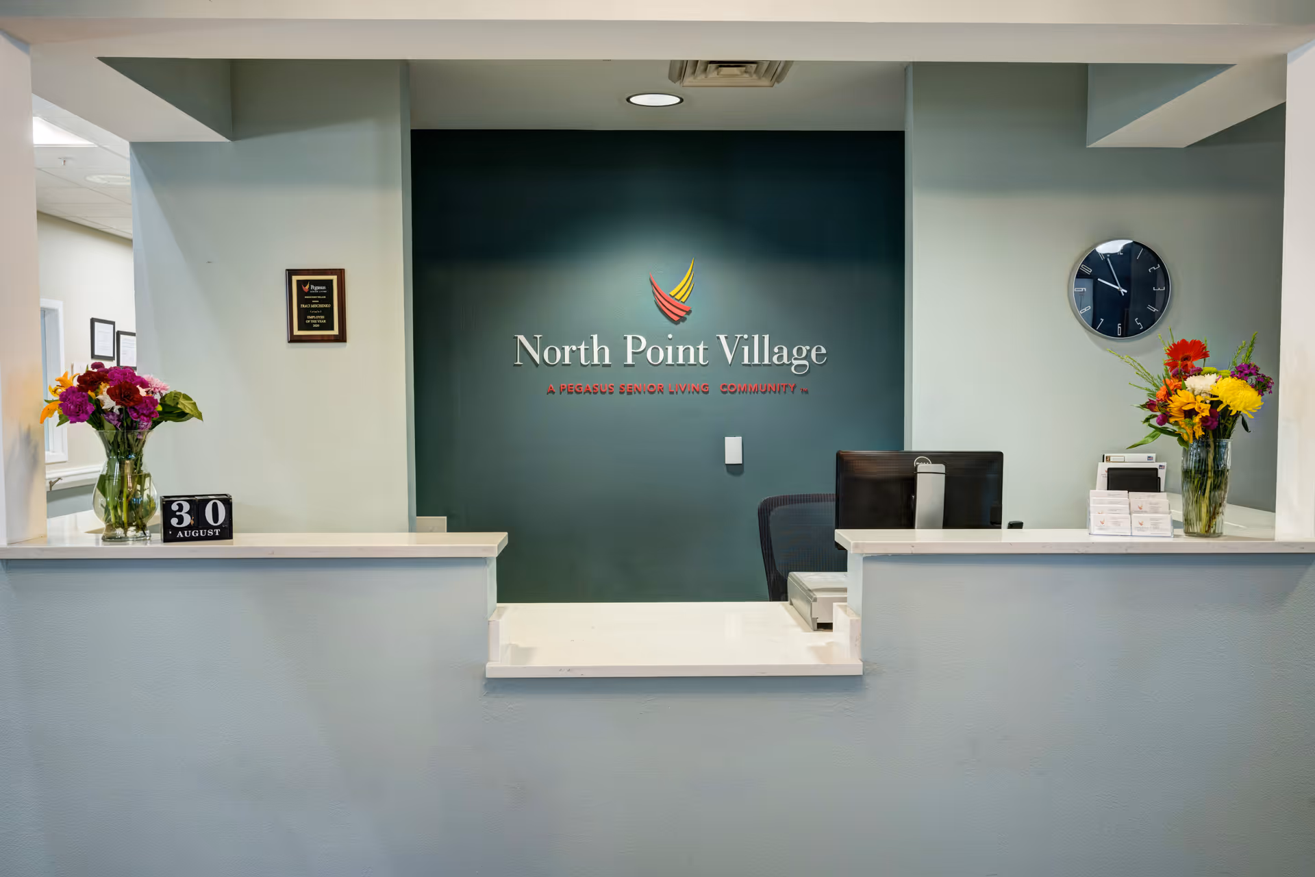 Reception desk area of North Point Village senior living community with a dark green accent wall displaying the facility name and logo. The desk has two vases with colorful flowers, a calendar showing August 30, a computer monitor, and a wall clock.